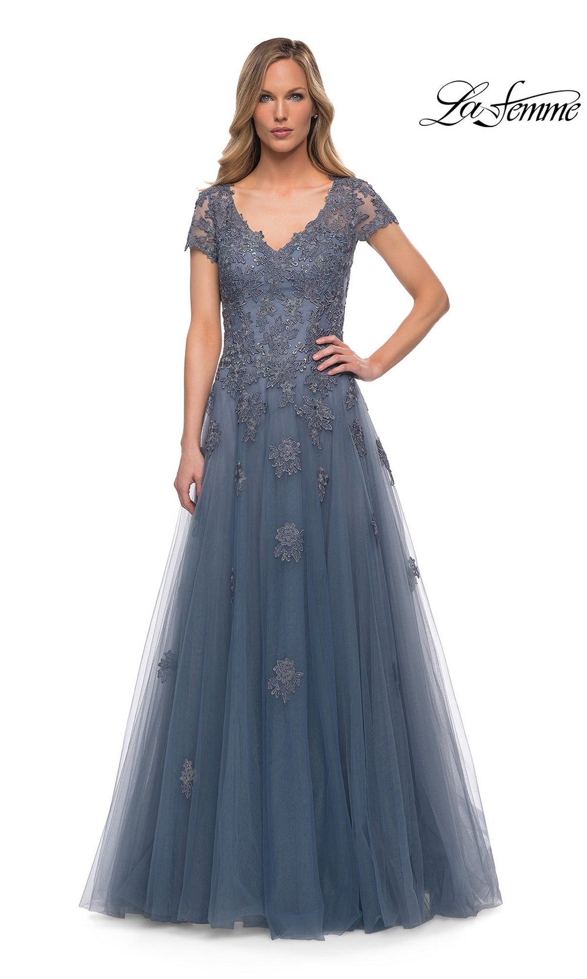 A woman in the Long Formal Dress: La Femme 29380, floor-length, short-sleeve, dusty blue with floral embroidery and lace appliques, stands against a white background.